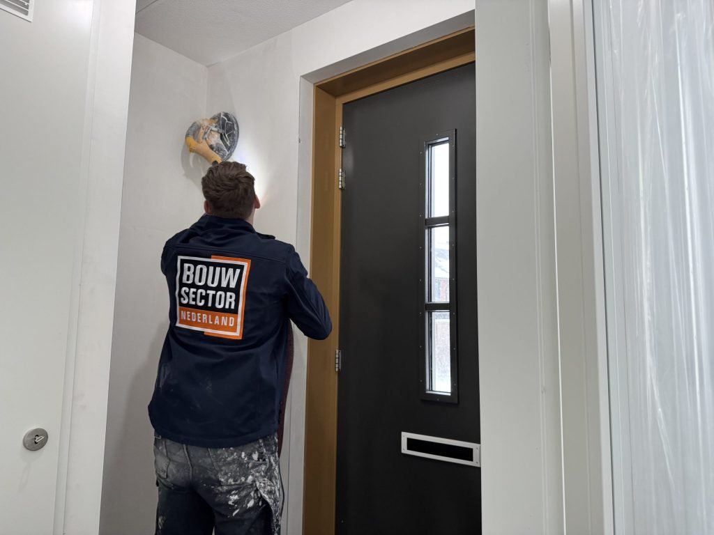 Stukadoor schuurt de aansluiting tussen wand en plafond in een hal naast de voordeur