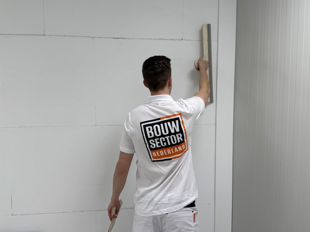 Stukadoor met het Bouwsector-logo op zijn rug strijkt een wand glad met een brede spaan