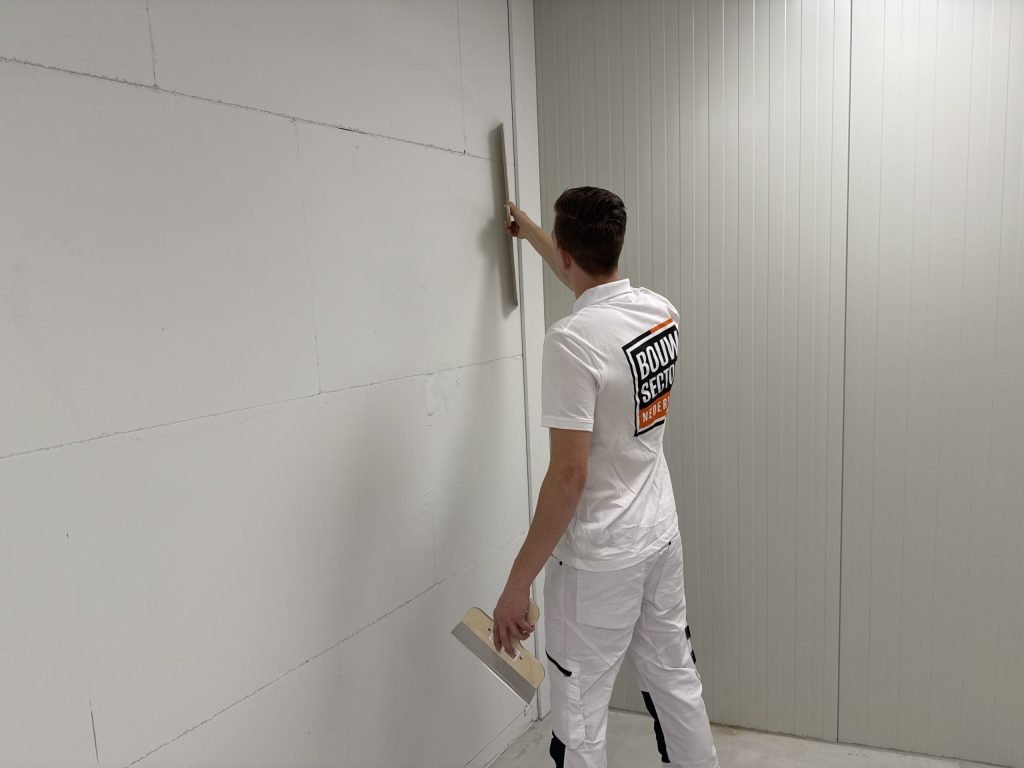 Stukadoor zet een lange spaan tegen een wand van witte platen in een lege ruimte