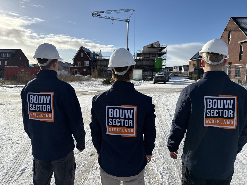 Drie vakmensen van Bouwsector Nederland met witte helm kijken naar nieuwe woningen en een bouwkraan in de sneeuw